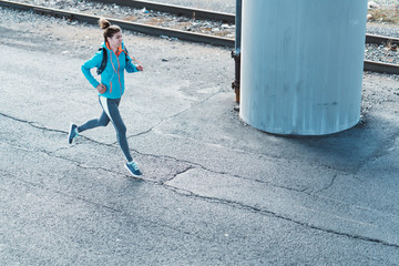 Urban sporty woman with water backpack and listening music running under the bridge in industrial city