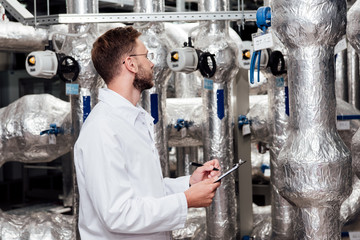 bearded engineer in white coat holding pen and clipboard near air compressed system