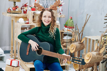 Portrait of young pretty smiling woman in reindeer antlers playing guitar and singing song