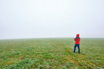 Rear view of man in red jacket standing on a green meadow and looking on his smartphone in front of a foggy nowhere landscape. Seen in October in Germany, Bavaria near Oedenberg.