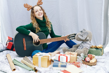 Portrait of happy young Caucasian woman sitting on the floor among giftboxes and toys and playing guitar