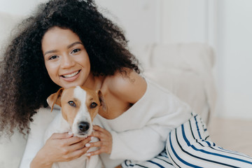 Image of attractive dark skinned woman wears makeup, has bushy curly hair, smiles pleasantly, cuddles dog, dressed in fashionable clothes, enjoys sweet moment with puppy, being at home together