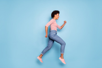Fototapeta na wymiar Side profile full length body size photo of cheerful positive cute pretty beautiful girlfriend wearing jeans denim striped shirt footwear running jumping isolated pastel blue color background