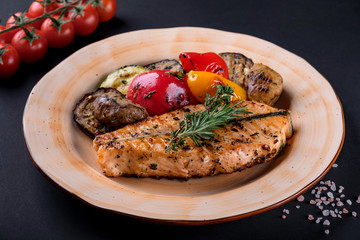 Top view grilled steak salmon fillet with vegetable salad, pepper, herbs on plate on black background. Delicious fried fish served coarse salt and cherry tomato branch for dinner. Healthy eat