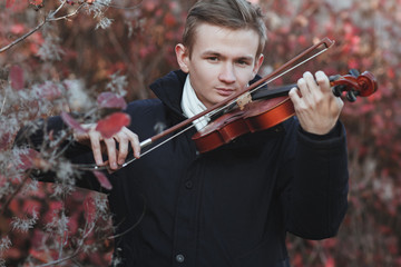portrait of a young elegant man playing the violin on autumn nature backgroung, a boy with a bowed...