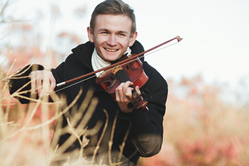 Fototapeta premium portrait of a young elegant violinist playing the violin in autumn field in grass, young man engaged in musical art, concept of classical music, hobby and relax