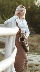 portrait of a beautiful young woman standing and leaninf on wooden white fence with saxophone,...