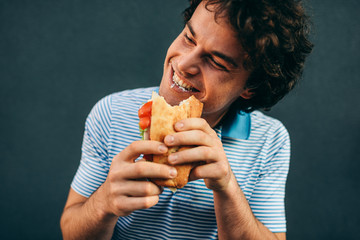 Portrait of young man eating a cheeseburger has pleasant expression. Happy man in a fast food restaurant eating a hamburger outdoors. Man with curly hair having street food and eat a burger.