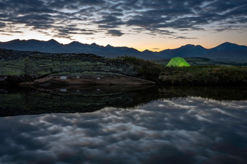 Green tent lighted up in the dark. Sunset and golden horizon in the back together with the mountain chain of Rondane in Norway. landscape, nature, camping, tent, hiking, sleep, reflection concept.