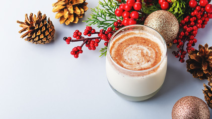 Traditional Christmas cocktail Eggnog with eggs, alcohol, grated nutmeg and cinnamon closeup. Sweet traditional drink on grey table with beige decorations, burning lanterns and pine cones