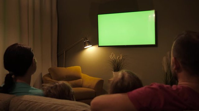 Family Sitting Together Sofa In Their Living Room Night Watching TV Green Screen. Rear View Of Family With Children Sitting On Sofa In Living Room Evening Watching Green Mock-up Screen TV Together.