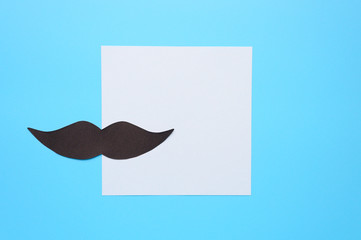 Black paper moustache and writing paper sticker on a blue background. The concept of awareness of men's health and prostate cancer. Space for text