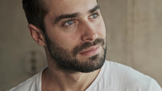 Close up view of thougthful handsome young brunet man smiling and touching his beard while looking to the side at home