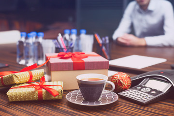 Young businessman surrounded by gifts in office. Workplace, cup of tea, laptop, ip phone and boxes. Christmas gift from colleague. Secret Santa office tradition. New year background.