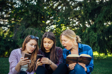 Three cute girls relax and socialize on the lawn in the summer park. Young women on the green grass among the trees, looking at smartphone, reading a book. Students in between classes outdoor.
