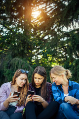 Three cute girls relax and socialize on the lawn in summer park. Young women sit on the green grass among the trees and look into the smartphone. Students in between classes, vertical photo, close up.