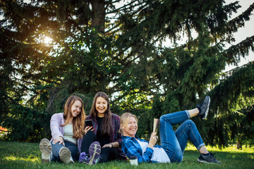 Happy young girls have fun laughing sitting on green grass in the park. Students in between lectures. Blonde, brunette and red-hair girl communicate, look at smartphone, concept of female friendship.