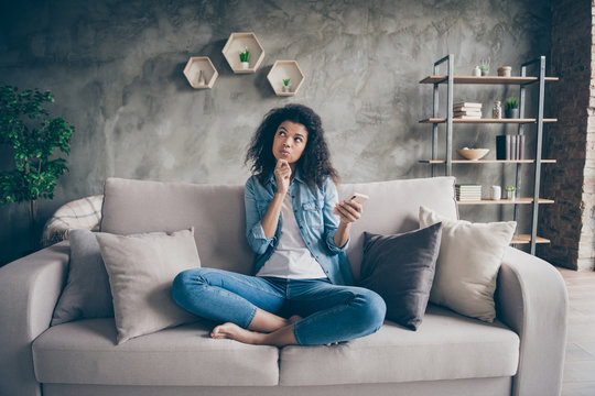 Photo of pretty dark skin curly lady hold telephone hand think over new port text finger on chin have doubts sitting comfy couch casual denim outfit living room indoors