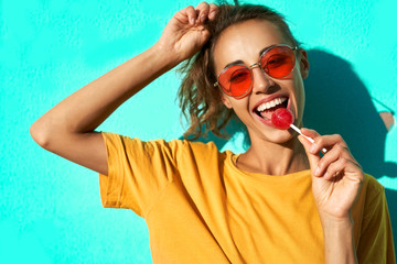 portrait of a young stylish woman smiling at camera, licking big red lollipop against blue background. - obrazy, fototapety, plakaty
