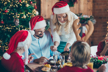 Photo of cheerful positive cute beautiful wife caring about her husband in santa hat headwear with their son wearing antlers