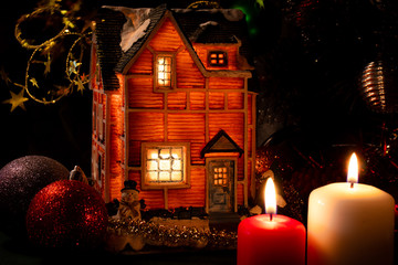 Christmas toy house-lamp stands under a Christmas tree with balls next to candles