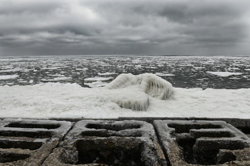 Gloomy dramatic winter sea with frozen water. Concrete structures ashore covered with ice and snow.