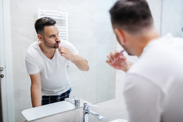 Handsome caucasian man standing in bathroom in pajamas and washing his teeth in the morning and watching himself in the mirror.