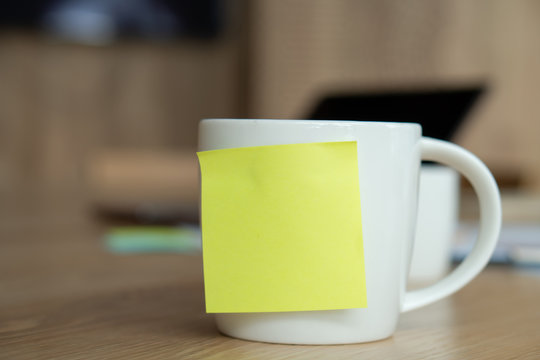 A white coffee cup with a blank yellow post-it on the cup. Resting on the desk and laptop is placed on the back.