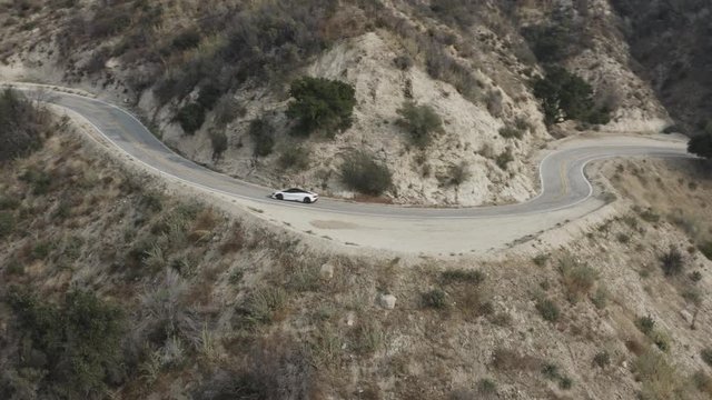 Aerial shot of a fast, white car taking a sharp turn on the windy interstate roads found in the desert hills of California.