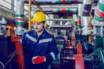Dedicated handsome caucasian blue collar worker in protective suit, with hardhat and antiphons checking on machine while standing in factory.
