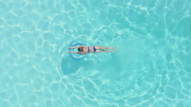 A woman in ablue bikini swimming in a pool with an inflatable ring. Aerial zoom out from directly above.