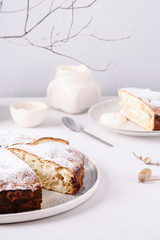 Homemade simple apple cake or pie sliced and sprinkled powdered sugar on grey concrete table decorated dry twig in vase. Autumn or winter treat. Fika. Hygge