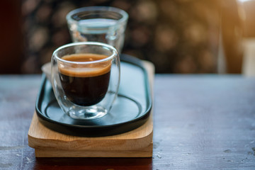 Hot espresso coffee in a clear glass, placed on a metal tray and wooden layer beside the coffee cup...