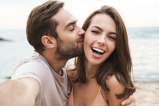 Image of young happy man kissing and hugging woman while taking selfie