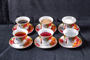 Six cups and saucers filled with water, juice, tea, coffee, milk and coffee with milk on a dark fabric background
