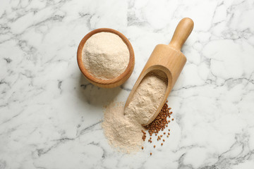 Bowl and scoop of buckwheat flour with grains on marble table, flat lay