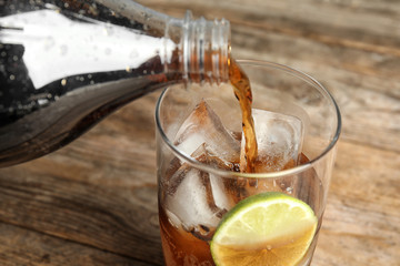 Pouring refreshing soda drink into glass with ice cubes and lime on wooden table, closeup