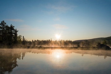 Beautiful morning orange sunrise over lake Sjabatjakjaure with haze mist in Sweden Lapland nature. Mountains, birch trees, spruce forest, rock boulders and grass. Sky, clouds and clear water.