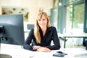 Portrai tof middle aged businesswoman sitting at office desk while looking at camera and smiling - Powered by Adobe