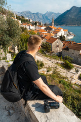 Fototapeta na wymiar Man traveler with backpack looking at old european town from the top of hill and sitting near photo camera