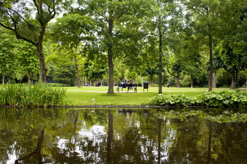 Fototapeta na wymiar View of a couple sitting on a bench, people hanging out, trees, grass field and pond at Vondelpark in Amsterdam. It is a public urban park of 47 hectares. It is a summer day.