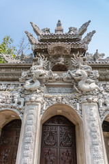 Fototapeta na wymiar Da Nang, Vietnam - March 10, 2019: Chua An Long Chinese Buddhist Temple. Fisheye closeup on Gray stone Pagoda entrance with brown doors and roof structure under light blue sky and with green foliage.