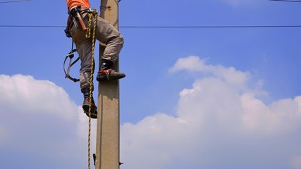 Low section of electrical lineman with tools and equipment is climbing on electric power pole against white cloud and blue sky background