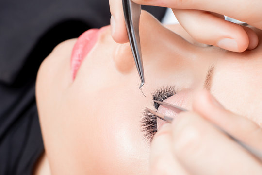 Eyelash extension procedure. Master tweezers sets fake lashes on beautiful woman