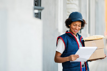 Happy African American mailwoman doing through check list while delivering packages.