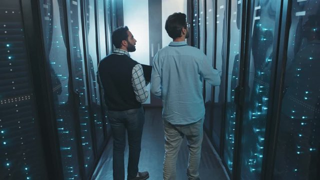 Two male server specialists walking in server room collaborating examining rack data cabinets working behind glass panels. Data center. Teamwork.