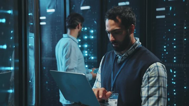 Caucasian IT technician webhosting in digital server room looking into data cabinets typing on computer laptop. Data center. Technology and people.