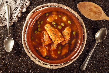 Closeup of a clay pot with a delicious fish stew with peas and potatoes cooked with spoons and vintage ornaments on a stone table. Healthy fish soup. Homemade food. Hot food, freshly made