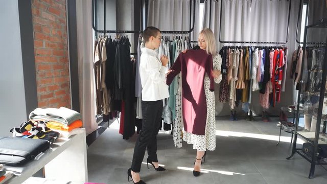 Beautiful caucasian woman with blond hair in white dress holding new collection clothing by fashion designer. Background rack of clothes in studio