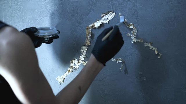 Woman in black protective gloves is applying golden paint to the grey wall with small brush.